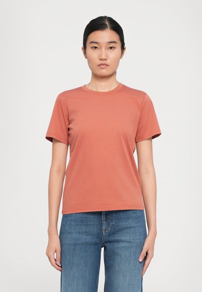 Mujer con cabello oscuro recogido, vistiendo una camiseta de manga corta de color óxido liso y jeans azules, frente a un fondo blanco.