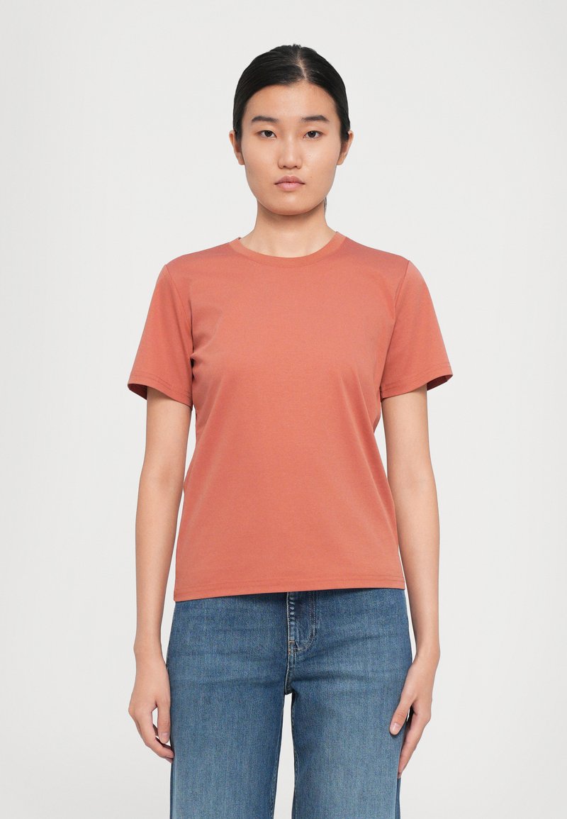Kvinna med mörkt hår som är uppsatt, iförd en enfärgad rostfärgad t-shirt med korta ärmar och blåjeans mot en vit bakgrund.
