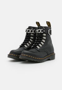 Černé kožené kotníkové boty se stříbrným řetízkem, kovovými očky a žlutým prošíváním na podrážce. Obsahují poutko na patě.