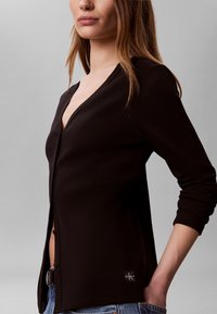 Жена с черен кардиган с копчета с лого на Calvin Klein от долната страна и сини дънки, стояща с ръка в джоба.