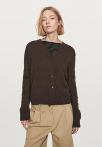 Brauner gestrickter Cardigan mit V-Ausschnitt, Knopfleiste und geripptem Saum. Getragen über einem schwarz-grün gestreiften Oberteil, kombiniert mit beigen Hosen.