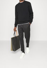 Felpa nera con collo rotondo, pantaloni grigi, sneakers bianche e una grande borsa a tracolla nera con manici beige. Texture liscia e vestibilità comoda.