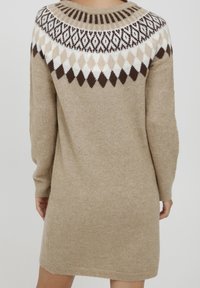 Strikket sweaterkjole i lys beige med et geometrisk mønster i brun, creme og hvid. Har lange ærmer og en rund halsudskæring.