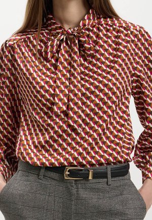 Femme portant un chemisier à motif géométrique avec un col lavallière, rentré dans un pantalon gris à carreaux avec une ceinture noire et une boucle dorée.