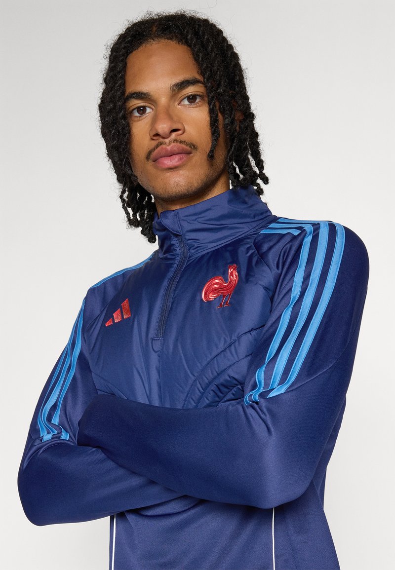 Veste de survêtement navy avec col montant, rayures bleues et logo de coq brodé en rouge. Elle comprend des manches longues et une fermeture éclair sur le devant.