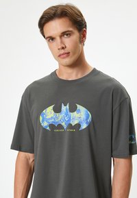 Camiseta de algodón gris con un gran logo azul de Batman que incluye ilustraciones de personajes y el texto "FOREVER BATMAN". Manga corta, corte casual.