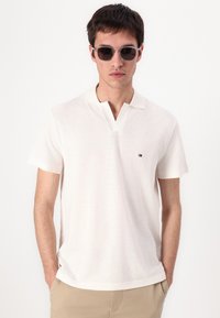 Polo a maniche corte color crema con scollo a V e un piccolo logo ricamato. Il modello indossa occhiali da sole grigi e pantaloni beige chiaro.