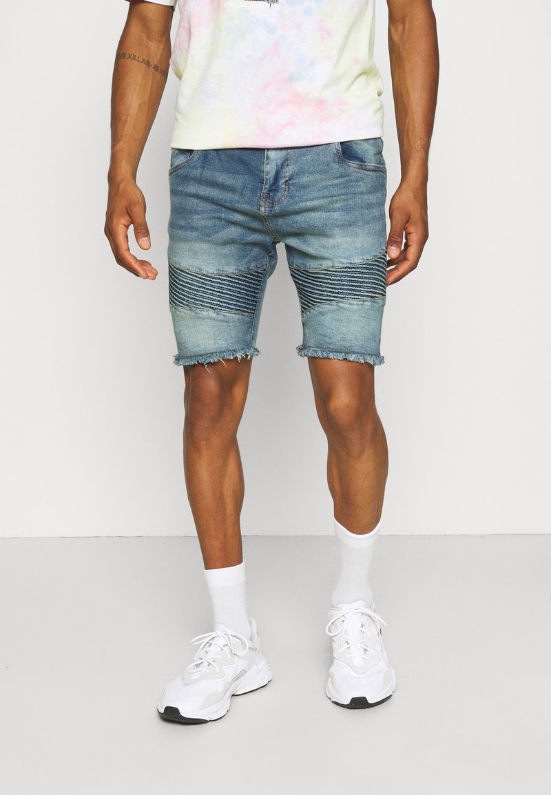 Homme portant un short en jean bleu délavé avec un détail de panneau côtelé et des ourlets effilochés, des chaussettes blanches, des baskets blanches et un t-shirt tie-dye pastel.
