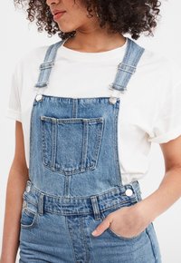 Pantalones de denim con tirantes ajustables, un bolsillo frontal y detalles en metal. Combinados con una camiseta de algodón blanca. Color azul claro.