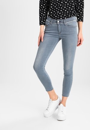 Femme portant un jean skinny bleu clair et des baskets blanches, debout avec les jambes croisées, portant un haut noir à manches longues avec des motifs triangulaires blancs.