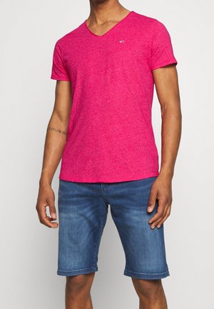 Mężczyzna ubrany w jasnoróżowy T-shirt z krótkim rękawem i dekoltem w serek oraz niebieskie dżinsowe szorty, stojący na jednolitym tle.