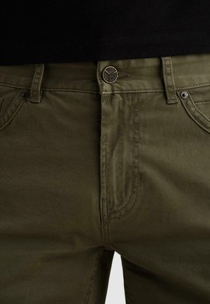 Zbliżenie na oliwkowozielone spodnie zapinane na metalowy guzik i suwak, noszone z czarną koszulą włożoną do środka.