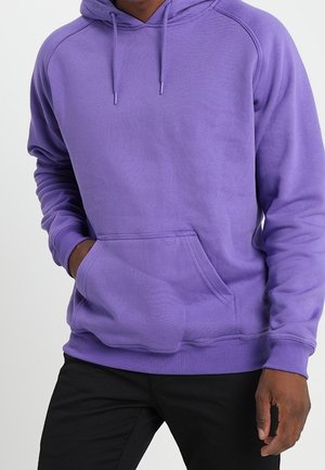 Mann trägt einen lila Hoodie mit Fronttasche und Kordeln, kombiniert mit schwarzen Hosen, steht vor einem einfarbigen Hintergrund.