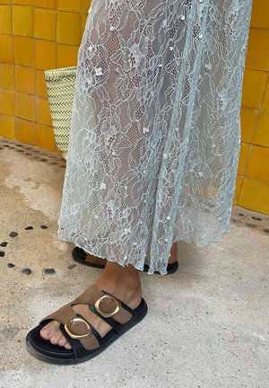 Piedi con sandali neri e marroni con grandi fibbie dorate, parzialmente coperti da una lunga gonna in pizzo azzurro pallido, in piedi su un pavimento in cemento testurizzato.