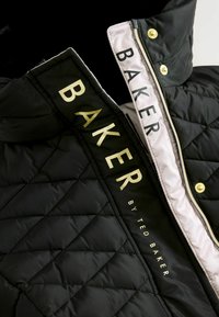 Veste matelassée noire avec lettre dorée "BAKER" et "by TED BAKER" sur un intérieur clair contrastant. Accents en métal doré.
