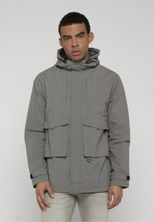 Mężczyzna w szarej kurtce z kapturem, z przednimi kieszeniami z klapami i regulowanymi mankietami, stojący na jasnym, jednolitym tle.