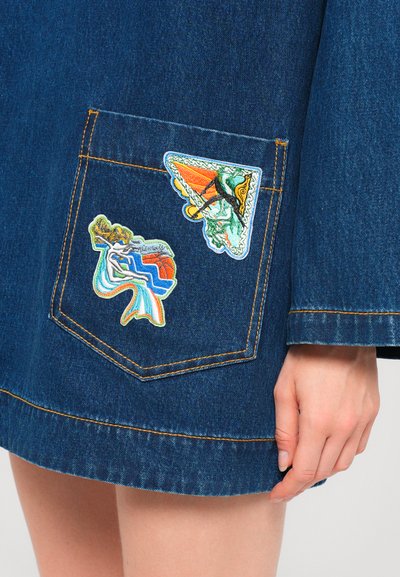Denim skirt with a dark blue wash, featuring three colorful embroidered patches on the pocket. Contrast stitching along seams and hem.