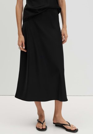 Femme portant une jupe midi noire, un haut noir sans manches, des sandales flip-flop à talon noires et des bagues argentées à la main gauche.