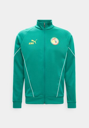 Grüne Sportjacke mit durchgehendem Reißverschluss, gelbem Puma-Logo und dem Schriftzug „KING“ rechts, Emblem der senegalesischen Fußballnationalmannschaft auf der linken Brust, weiße Paspelierung.