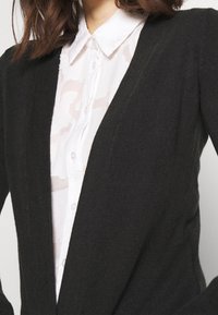 Czarny kardigan wykonany z miękkiego materiału, z dekoltem w serek i dopasowanym krojem, noszony na białej koszuli zapinanej na guziki.