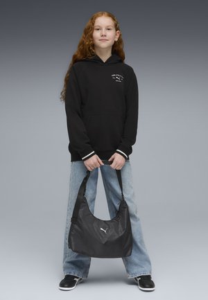 Jeune personne aux longs cheveux roux portant un sweat à capuche noir et un jean, tenant un sac à bandoulière noir Puma des deux mains devant.