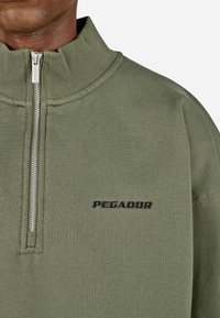 Zaļa flīsa džemperis ar augstu apkakli, priekšējo rāvējslēdzēju un ribota tekstūra. Melns "PEGADOR" logo kreisajā krūtī.