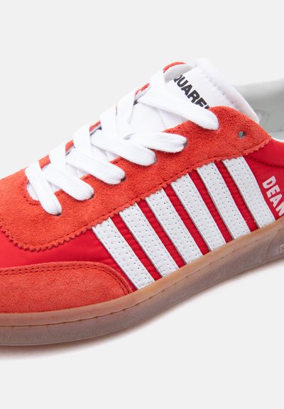Baskets basses rouges et blanches avec détails en daim, lacets blancs, bandes verticales blanches et semelle gomme, avec le texte « DEAN » sur le côté.