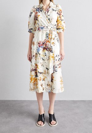 Abito a wrap floreale color crema con stampe colorate, maniche a sbuffo corte e cintura legata in vita. Presenta un orlo di lunghezza media. Sandali neri da infilare.