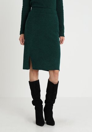 Femme portant une jupe vert foncé au genou avec une fente latérale et des bottes souples en daim noir à talons hauts, debout devant un fond uni.
