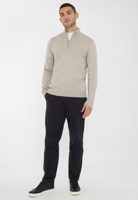 Beige zip-neck pullover in a smooth knit material, paired with dark pants and black sneakers, featuring a subtle logo detail at the hem.