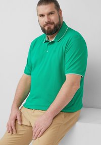 Chemise polo verte avec un accent blanc sur le col et les manches ; manches courtes, texture lisse ; portée par une personne assise devant un fond neutre.