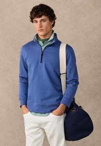 Jeune homme portant un sweat-shirt bleu à fermeture éclair quart, une chemise vert clair, un pantalon blanc, portant un sac marin bleu marine avec une sangle d'épaule beige.