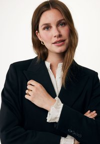 Svart kavaj med strukturerade axlar, bärs över en vit volangblus. Modellen har en guldring och creoleörhängen, poserar med armarna i kors.