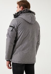 Geacă puffer gri cu glugă căptușită cu blană sintetică, buzunare laterale și manșete elastice. Are un material texturat și o potrivire lejeră.