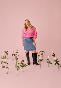 Femme en blouse rose, jupe en jean et bottes noires se tient parmi des vases contenant des roses rose clair sur un fond rose.