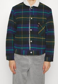 Chaqueta de cuadros en tonos negros, verdes, azules y amarillos. Cuenta con un cuello blanco y suave, botones a presión y un bolsillo en el pecho.