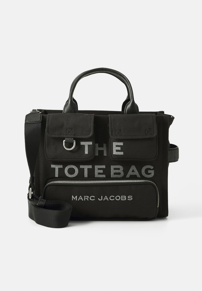 Sac en toile noir avec des accents en cuir, arborant un texte argenté "THE TOTE BAG" et "MARC JACOBS", des poignées doubles et une bandoulière amovible.