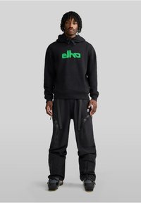 Felpa nera con logo verde, pantaloni neri con zip e cinghie, tessuto testurizzato, indossati con scarpe nere; design minimalista.