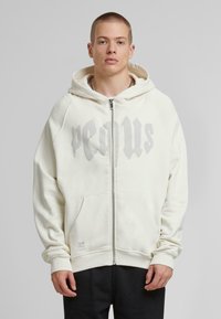 Cremefarbene Zip-Hoodie mit grauem Text, Kängurutaschen, gerippten Abschlüssen und Baumwollmaterial. Sanfte Textur und lässige Passform.