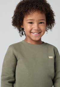 Maglione verde oliva con scollo rotondo e polsini a costine, dotato di un piccolo patch logo beige sulla parte sinistra del petto. Texture morbida.