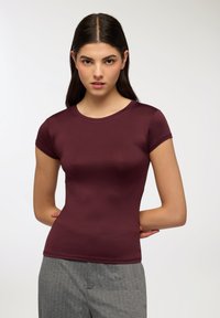 Top fitted a maniche corte color bordeaux realizzato in materiale liscio, con scollo rotondo e dettagli di cucitura sottili. Abbinato a pantaloni grigi a righe.