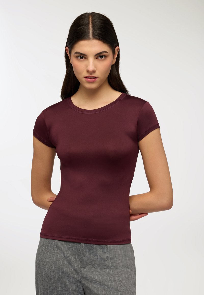 Top fitted a maniche corte color bordeaux realizzato in materiale liscio, con scollo rotondo e dettagli di cucitura sottili. Abbinato a pantaloni grigi a righe.