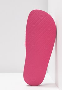 Semelle de sandale en caoutchouc rose avec des motifs de prise circulaire, présentée à la verticale contre un mur et un sol blancs dans un coin.