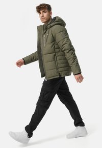 Gewatteerde groene jas met een capuchon, voorzien van een ritssluiting aan de voorkant en zijzakken, gedragen over zwarte cargobroeken en witte sneakers.