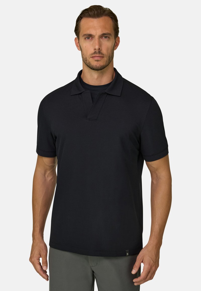 Homme aux cheveux bruns courts et légère barbe, portant un polo noir à manches courtes et un pantalon gris, debout devant un fond uni.