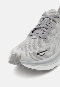 Scarpa da corsa grigia chiara con tomaia testurizzata, caratterizzata da un design in mesh e sistema di allacciatura. Suola bianca con ammortizzazione e un sottile dettaglio del logo.