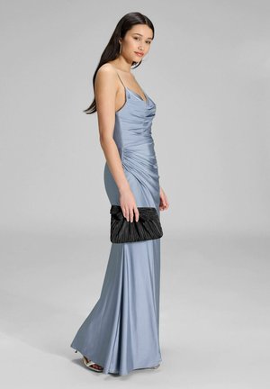 Eine Frau in einem hellblauen Satin-Abendkleid hält eine schwarze, plissierte Clutch und steht seitlich vor einem einfachen grauen Hintergrund.