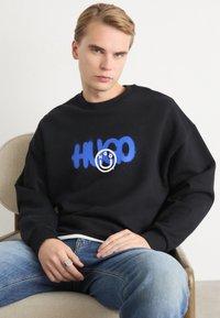 Crna dukserica s plavim sprejem napisanom "HUGO" i kružnom grafikom. Ima opušteni kroj i rebraste manžetne. Nosena je uz svijetloplave traperice.