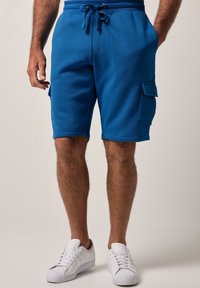 Pantaloni corti in misto cotone blu con vita regolabile tramite cordino, tasche laterali e tasca cargo sulla gamba destra. Abbinati a sneaker bianche.