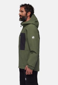 Veste imperméable verte avec une poche poitrine noire, une fermeture éclair, une capuche ajustable et un logo sur la manche gauche. Texture lisse.